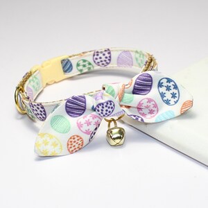 Puede incluir: Un collar blanco para gatos con un colorido estampado de huevos de Pascua. El collar tiene una hebilla de plástico amarilla y una campana dorada unida a una pajarita a juego. La pajarita presenta el mismo diseño de huevo.