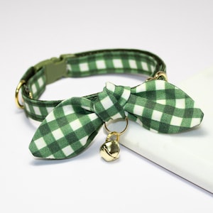 May include: Green and white gingham cat collar with a matching bow tie and a gold-colored bell. The collar has a green plastic buckle and gold-colored metal hardware. The bow tie is attached to the collar with a gold-colored ring.