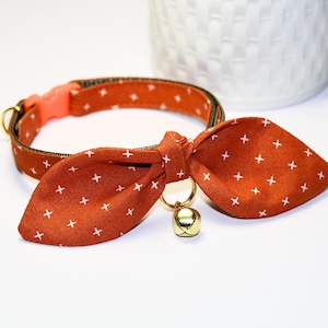 Puede incluir: Un collar de gato marrón y blanco con estampado, con una pajarita y una campanilla dorada. El collar es ajustable y tiene una hebilla de seguridad.