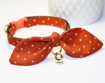 May include: A brown and white patterned cat collar with a bow tie and a gold bell. The collar is adjustable and has a safety buckle.