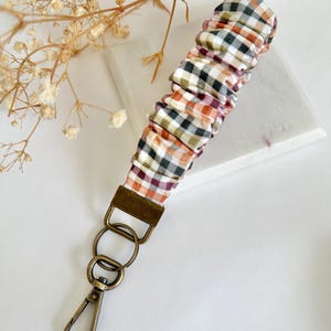 Puede incluir: Un llavero de pulsera de tela con un estampado de cuadros de temática otoñal en naranja, verde, blanco y burdeos. El llavero tiene un cierre de color bronce y anillas metálicas para sujetar las llaves. La pulsera mide unos 15 cm de largo.