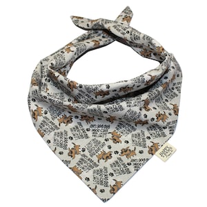 May include: A beige bandana with a brown dog print and the text "I can't talk right now, busy doing bad dog shit". The bandana is tied in a knot at the top and has a small brown tag with the text "Happy Paws" on it.