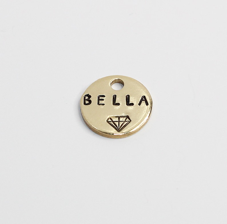 May include: Gold-colored metal tag with the word "BELLA" engraved in black lettering and a diamond symbol.