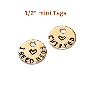 May include: Two gold-colored metal tags with a hole at the top. One tag says "I NEED MEDS" with a heart symbol. The other tag says "CHIPPED" with a heart symbol.