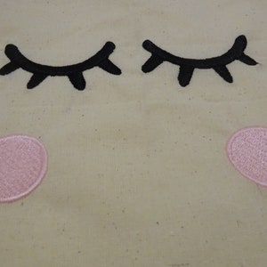 May include: A beige fabric with two black embroidered eyelashes and two pink embroidered circles.
