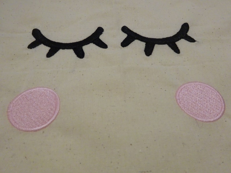May include: A beige fabric with two black embroidered eyelashes and two pink embroidered circles.