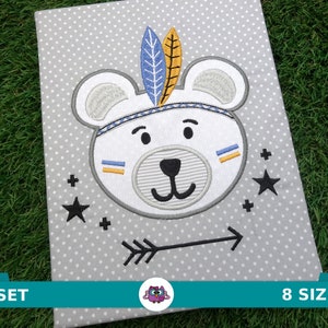 Puede incluir: Diseño bordado de un oso blanco con un tocado con una pluma azul y amarilla. El oso está rodeado de estrellas negras y signos más sobre un fondo gris. Una flecha está debajo del oso.