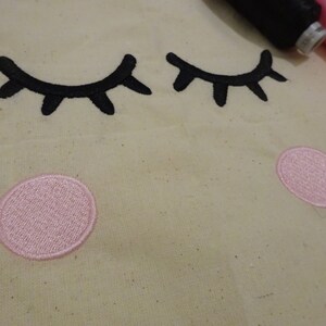 May include: A close-up of a beige fabric with two black embroidered eyelashes and two pink embroidered circles.
