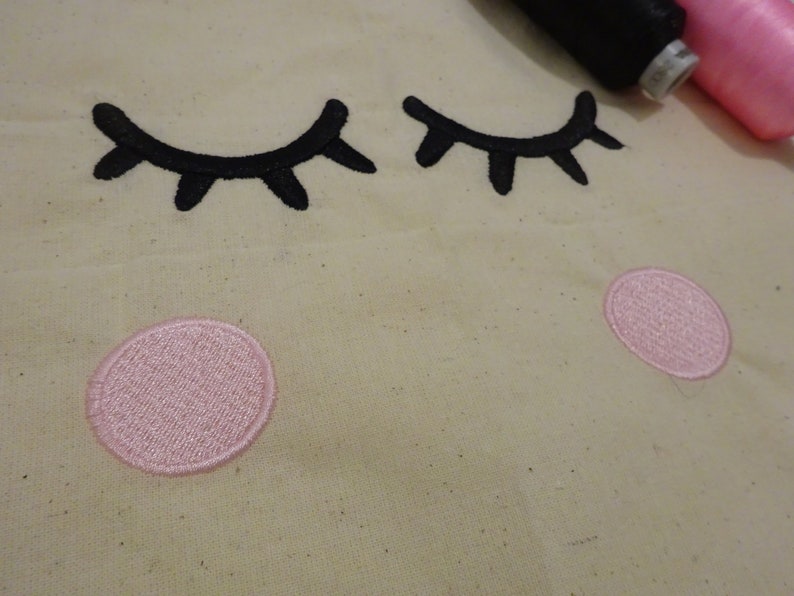 May include: A close-up of a beige fabric with two black embroidered eyelashes and two pink embroidered circles.