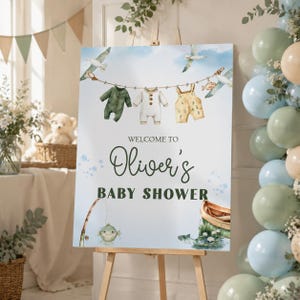 Könnte beinhalten: Ein rechteckiges Willkommensschild für eine Babyparty mit Aquarellillustrationen von Babykleidung auf einer Wäscheleine, Möwen, einer Angelrute, einem Fisch und einem kleinen Boot. Der Text lautet: WELCOME TO Oliver's BABY SHOWER in Dunkelgrün.