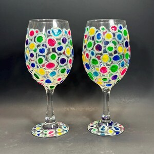 Può includere: Due calici da vino trasparenti decorati con pois colorati nelle tonalità di blu, verde, giallo, rosa e viola. I pois sono delineati con glitter argentati. I calici hanno stelo e base trasparenti.