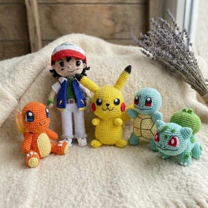 May include: A collection of handmade crochet Pokémon characters and Ash Ketchum dolls. The dolls are in various colors, including yellow, orange, blue, and green. The dolls are arranged on a cream-colored surface.