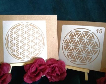 May include: Two square stencils featuring a white flower of life geometric pattern on a brown background. The right stencil displays the number 15 in the top right corner. Three dark pink artificial roses are positioned in front of the stencils.