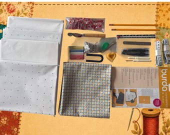 May include: A sewing supply kit featuring folded white dotted paper, a patterned fabric square, Burda tracing carbon paper, two yellow pencils, a metal ruler, measuring tapes, red clips, a seam ripper, pins, chalk, and a spool of red thread.