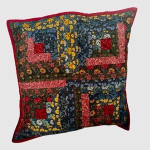 May include: A square throw cushion featuring a patchwork log cabin design. The fabric consists of various floral patterns in shades of red, yellow, blue, and dark green. The cushion is finished with a solid red fabric border along the outer edges.