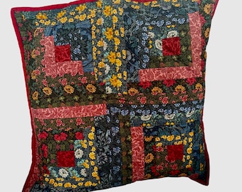 May include: A square throw pillow featuring a patchwork log cabin design. The fabric consists of various floral patterns in shades of red, yellow, blue, and dark green. The pillow is finished with a solid red fabric border along the outer edges.