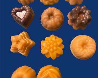 May include: A collection of eleven small, molded wax or soap pieces in various shapes including flowers, stars, hearts, and circular bundt-like forms. The items are primarily shades of orange, peach, and brown, arranged against a solid blue background.