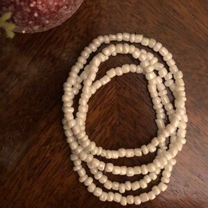 May include: A white beaded necklace with multiple strands. The necklace is made of small, round beads and is coiled up on a wooden surface.