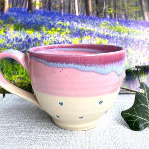 Könnte beinhalten: Handgefertigte Keramiktasse in Rosa und Creme. Der Rand ist rosa, mit einem hellblauen Wellenmuster, der Korpus ist rosa. Kleine blaue Herzen verzieren den cremefarbenen Boden. Die Tasse hat einen gebogenen Henkel. Fassungsvermögen ca. 350 ml.
