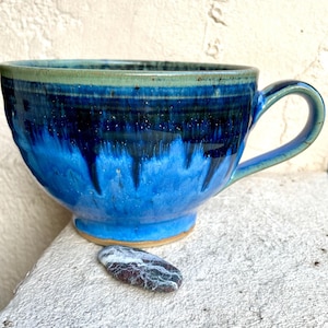 Könnte beinhalten: Eine blaue und grüne Keramiktasse mit einem gesprenkelten Überzug. Die Tasse hat eine breite, abgerundete Schale und einen Henkel. Ein kleiner, weißer und grauer Stein liegt vor der Tasse.