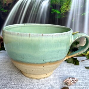 Könnte beinhalten: Eine handgefertigte Keramik-Tasse mit einer breiten, abgerundeten Form und einem großen Henkel. Die Tasse hat ein zweifarbiges Design, mit einer hellgrünen oberen Hälfte und einer gesprenkelten beige-farbenen unteren Hälfte. Geeignet für Kaffee, Tee oder Suppe.