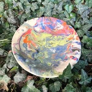 Könnte beinhalten: Eine runde Keramikschale mit einem marmorierten Oberflächendesign mit wirbelnden Mustern in hellem Limettengrün, leuchtendem Orange, tiefem Blau und Weiß. Das Innere der Schale zeigt eine glatte, glänzende Oberfläche mit vermischten Farben.