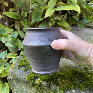 Könnte beinhalten: Ein handgefertigter, dunkelbrauner Keramiktopf mit einem Wellenlinienmuster am oberen Rand. Der Topf wird in einer Hand gehalten und steht auf einem moosbewachsenen Felsen, mit grünen Blättern im Hintergrund. Rustikale, erdige Ästhetik.