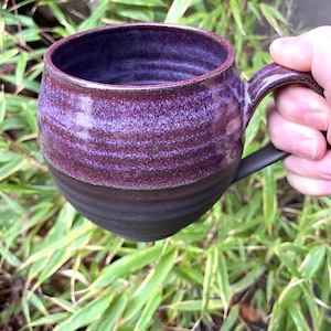 Könnte beinhalten: Handgefertigte Keramiktasse in Dunkelviolett und Schwarz. Der obere Teil der Tasse ist ein strukturiertes, leuchtendes Violett, das sich von der tiefschwarzen unteren Hälfte abhebt. Die Tasse hat eine runde Form und einen stabilen Henkel.