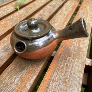 Könnte beinhalten: Eine kleine, braune und orangefarbene Teekanne aus Keramik mit Deckel und langem Ausguss. Die Teekanne hat eine gesprenkelte Glasur und eine raue Textur.