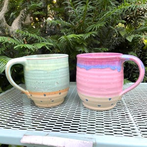 Könnte beinhalten: Zwei handgefertigte Keramikbecher. Einer ist hellgrün und beige mit orangefarbenen Akzenten. Der andere ist rosa mit blauen und violetten Akzenten. Beide Becher haben einen Henkel und stehen auf einer weißen Metalloberfläche.