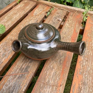Könnte beinhalten: Eine kleine, dunkelbraune Teekanne aus Keramik mit Deckel und Ausguss. Die Teekanne hat eine gesprenkelte Glasur und eine leicht abgerundete Form.