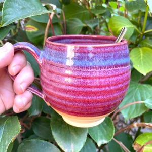 Könnte beinhalten: Ein runder Keramikbecher mit gerippter Textur, der einen lebendigen Farbverlauf aus tiefem Magenta und violetter Glasur aufweist, welcher in einen natürlichen beigen Boden übergeht. Der Becher hat einen geschwungenen Henkel und einen silbernen Löffel.