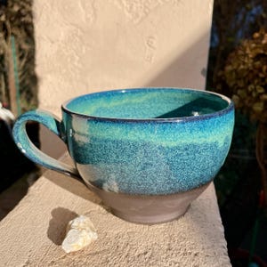 Könnte beinhalten: Handgefertigte Keramiktasse mit türkisfarbener und blaugrüner Glasur. Die Tasse hat einen dunkelbraunen Boden und einen gebogenen Henkel. Die Tasse steht auf einer Steinoberfläche. Ideal für Tee, Kaffee oder andere Heißgetränke.