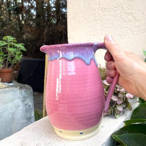 Könnte beinhalten: Ein Keramikkrug mit einem glänzend rosa Körper und einer glatten, hellblauen Tropfglasur am Rand. Der Boden ist cremefarben und mit kleinen, gleichmäßig verteilten blauen Herzakzenten versehen. Er hat einen geschwungenen Henkel und einen Ausguss.
