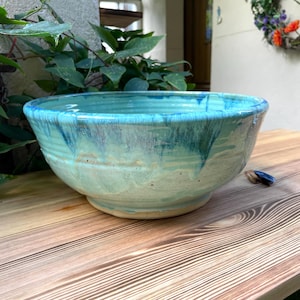 Könnte beinhalten: Eine handgefertigte Keramikschale mit einem breiten, flachen Design. Die Schale hat eine türkisfarbene Glasur mit dunkleren blauen Tropfen, die einen geschichteten Effekt erzeugen. Die Schale steht auf einer Holzoberfläche und zeigt ihre einzigartigen, künstlerischen Qualitäten.