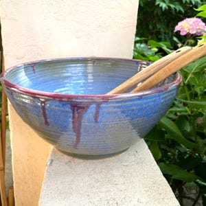 Könnte beinhalten: Eine große, runde Keramikschale mit blauer Glasur und sichtbaren horizontalen Rillen. Der Rand weist ein dunkelviolettes Tropfmuster auf, das an den Außenseiten herabläuft. Zwei hellbraune hölzerne Salatbesteckteile liegen in der Schale.