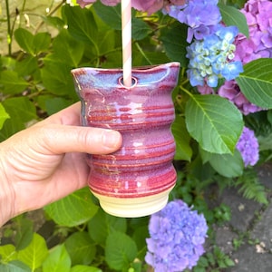 Könnte beinhalten: Ein Keramikbecher mit glänzender, mehrfarbiger Oberfläche in Schattierungen von tiefem Magenta, Violett und Blau. Das Gefäß weist eine wellenförmige, gerippte Textur am Körper und eine kleine kreisförmige Öffnung am oberen Rand auf.
