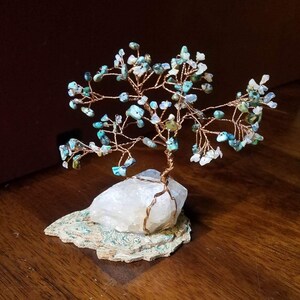 May include: A wire-wrapped crystal tree with turquoise and white stones on a clear quartz base. The tree is on a wooden surface.