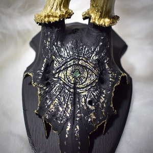 May include: A black and gold deer skull with a green gemstone eye painted on the front. The skull is mounted on a black wooden plaque.
