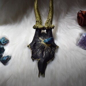 May include: A black deer skull with gold antlers and a teal blue crystal embedded in the top. The skull is surrounded by a white fluffy surface and other crystals.