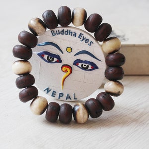 May include: A brown and white beaded bracelet with a round charm featuring a painted face with blue eyes and the text "Buddha Eyes Nepal".
