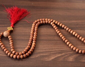 May include: A wooden mala bead necklace with a red tassel and a decorative bead. The beads are light brown and round, strung together to form a necklace. The tassel is bright red and attached to the necklace.