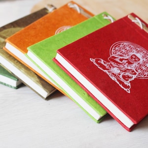 May include: A stack of colorful handmade journals with white pages and stitched spines. The covers are in shades of green, brown, orange, and red. The red journal features a white illustration of a seated figure.