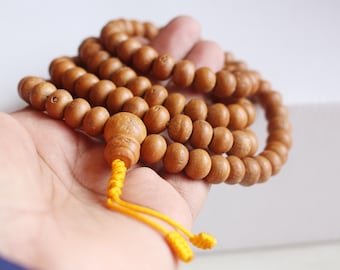 May include: A strand of prayer beads made of light brown wooden beads. The beads are held in a hand, with a yellow tassel at the end. The beads are round and smooth, with a natural wood grain.
