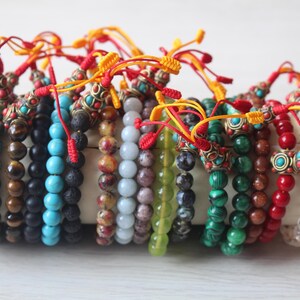Puede incluir: Una colección de pulseras de cuentas coloridas con detalles metálicos decorativos y lazos de cuerda roja y amarilla. Las pulseras están hechas de varias piedras naturales, incluyendo turquesa, ojo de tigre y roca volcánica.