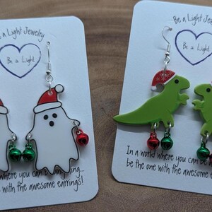 May include: Two pairs of Christmas-themed earrings. One pair features white ghosts wearing Santa hats with green and red jingle bells. The other pair features green dinosaurs wearing Santa hats with green and red jingle bells. The cards have the text "Be a Light Jewelry" and "In a world where you can be anything... be the one with the awesome earrings!"
