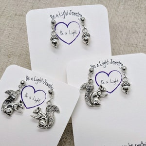 May include: Three pairs of silver earrings with a squirrel and acorn charm. The earrings are displayed on white cards with the text "Be a Light Jewelry" and "Be a Light".