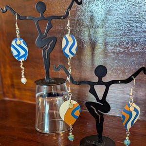 Puede incluir: Un par de pendientes colgantes con patrones abstractos azules, blancos y naranjas en la parte delantera. Los pendientes cuelgan de una cadena plateada y tienen una pequeña perla y una cuenta azul y verde en la parte inferior.