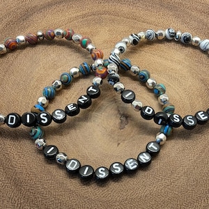 Puede incluir: Tres pulseras de cuentas con la palabra "DISSENT" escrita en letras blancas sobre cuentas negras. Las pulseras presentan una mezcla de cuentas coloridas y plateadas, sobre un fondo de madera.