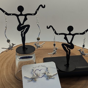 May include: Silver-toned roadrunner earrings with dangling beads and a curved silver accent. The earrings are displayed on a black stand, with a pair of stud earrings on a white card that reads "Be a Light Jewelry".